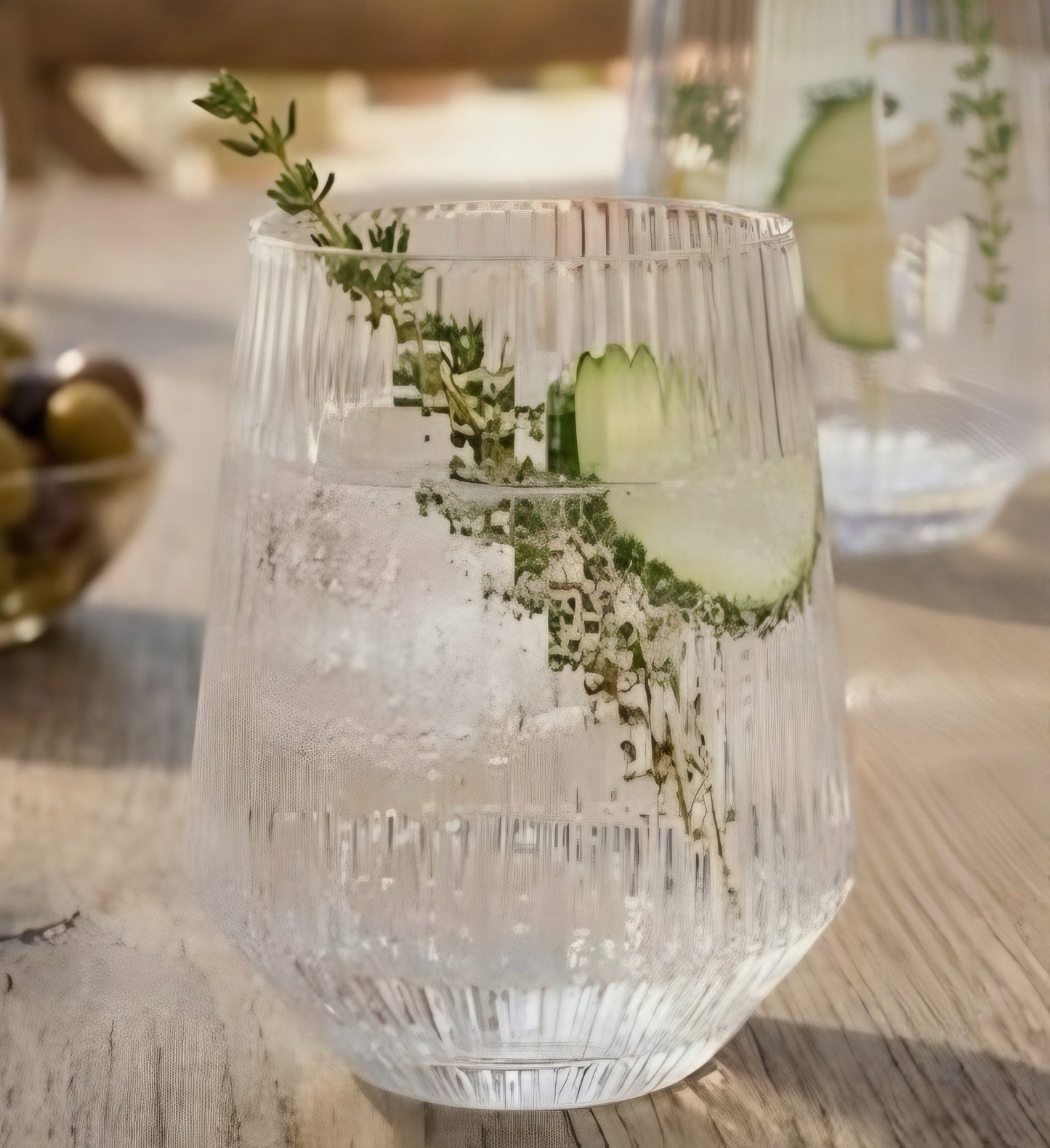 Bicchiere in vetro trasparente con texture rigata plissé, riempito con ghiaccio e guarnizione di cetriolo, poggiato su un tavolo in legno in stile lifestyle.