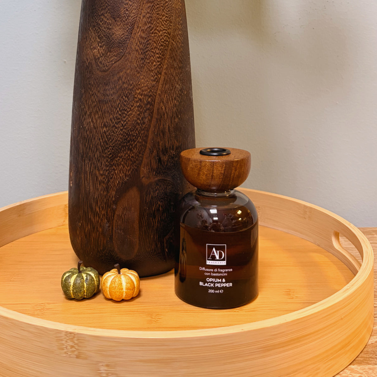 Diffusore profuma ambienti Opium & Black Pepper in flacone di vetro ambrato con tappo in legno, ambientato su vassoio con vaso in legno e zucche decorative autunnali