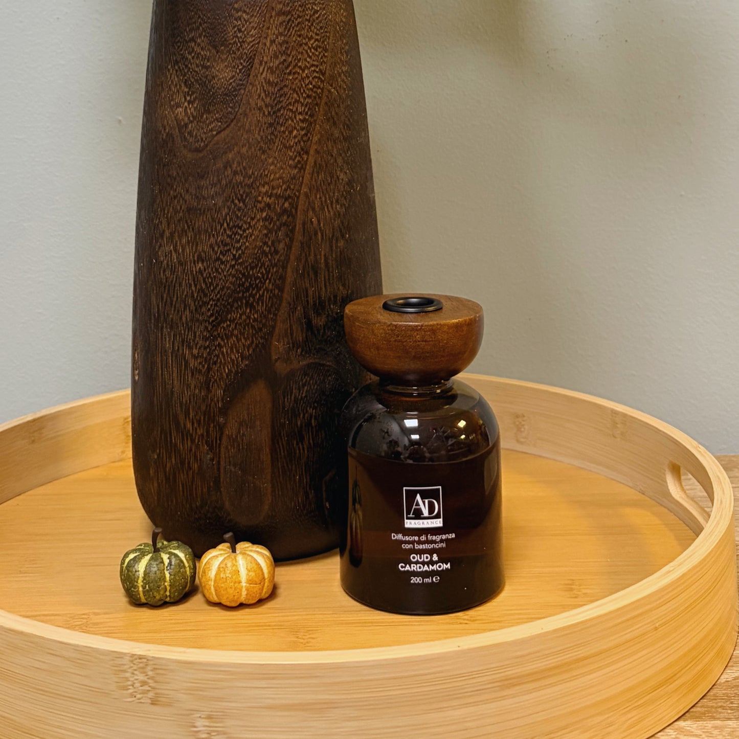 Diffusore profuma ambienti Oud & Cardamom in flacone di vetro ambrato con tappo in legno, ambientato su vassoio con vaso in legno e zucche decorative autunnali
