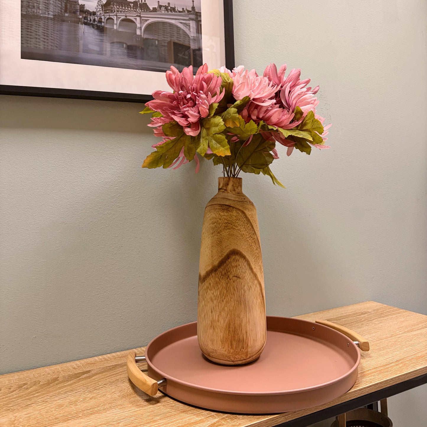 Composizione floreale con fiori finti rosa all'interno di un vaso in legno chiaro, appoggiato su un vassoio di design rosa antico sopra una console di legno.