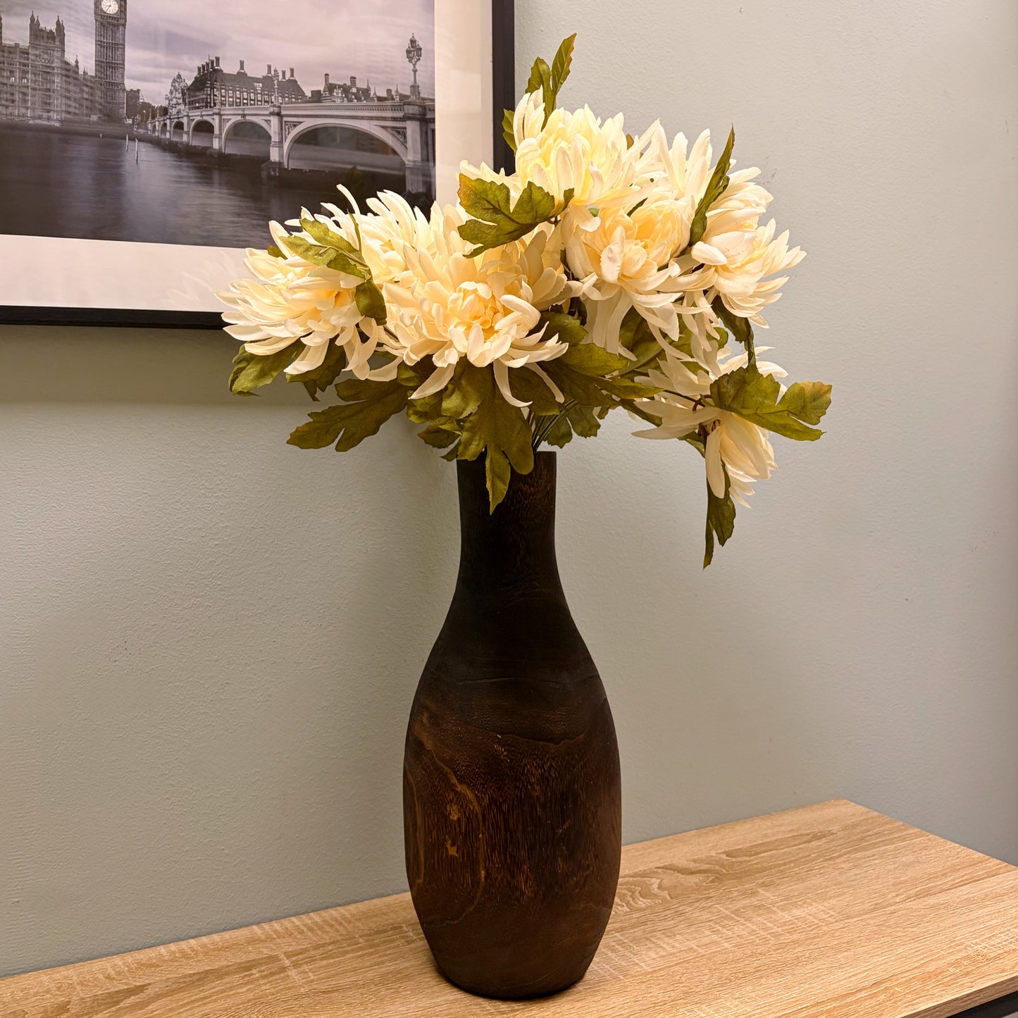 Vaso alto in legno scuro con bouquet di fiori finti bianchi posizionato su un tavolo in legno davanti a una parete salvia.