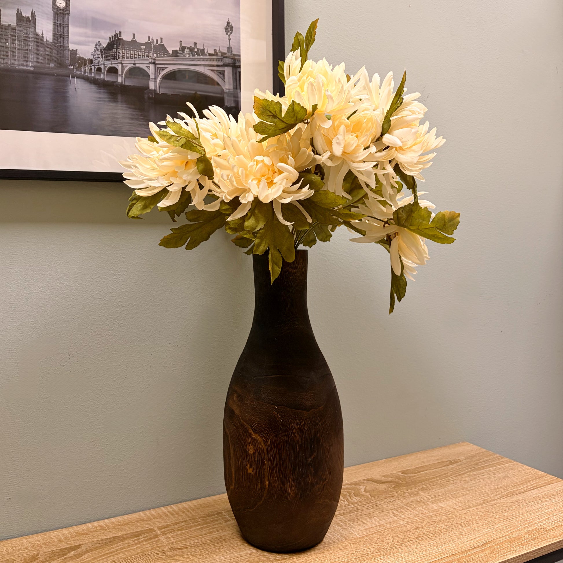 Vaso alto in legno scuro con bouquet di fiori finti bianchi posizionato su un tavolo in legno davanti a una parete salvia.