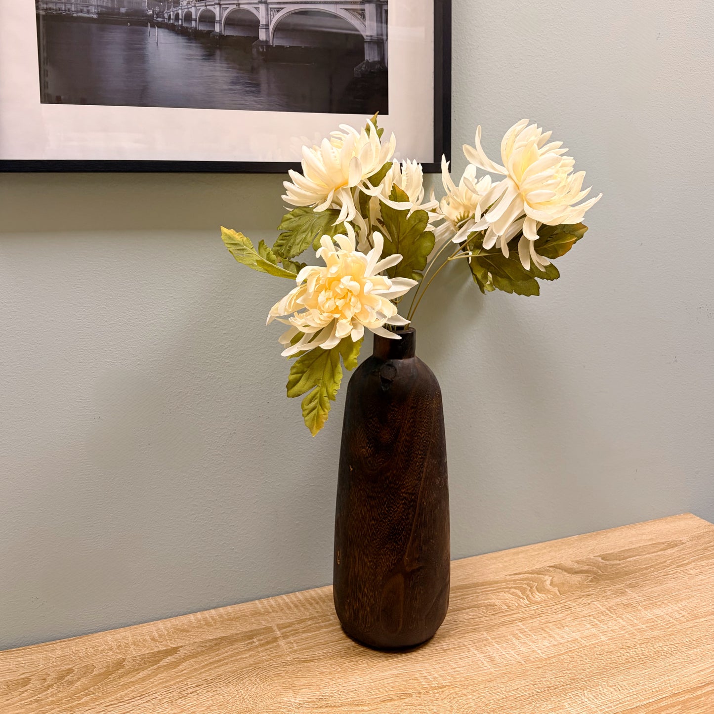 Vaso alto in legno scuro con bouquet di fiori finti bianchi posizionato su un tavolo in legno davanti a una parete salvia.