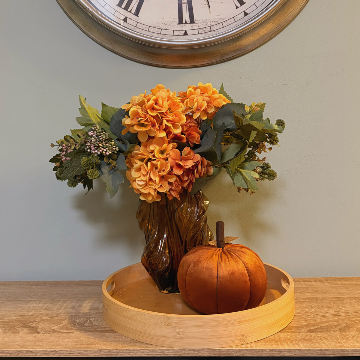 Zucca decorativa in velluto colore arancio caramello ambientata su vassoio in legno con vaso e fiori autunnali