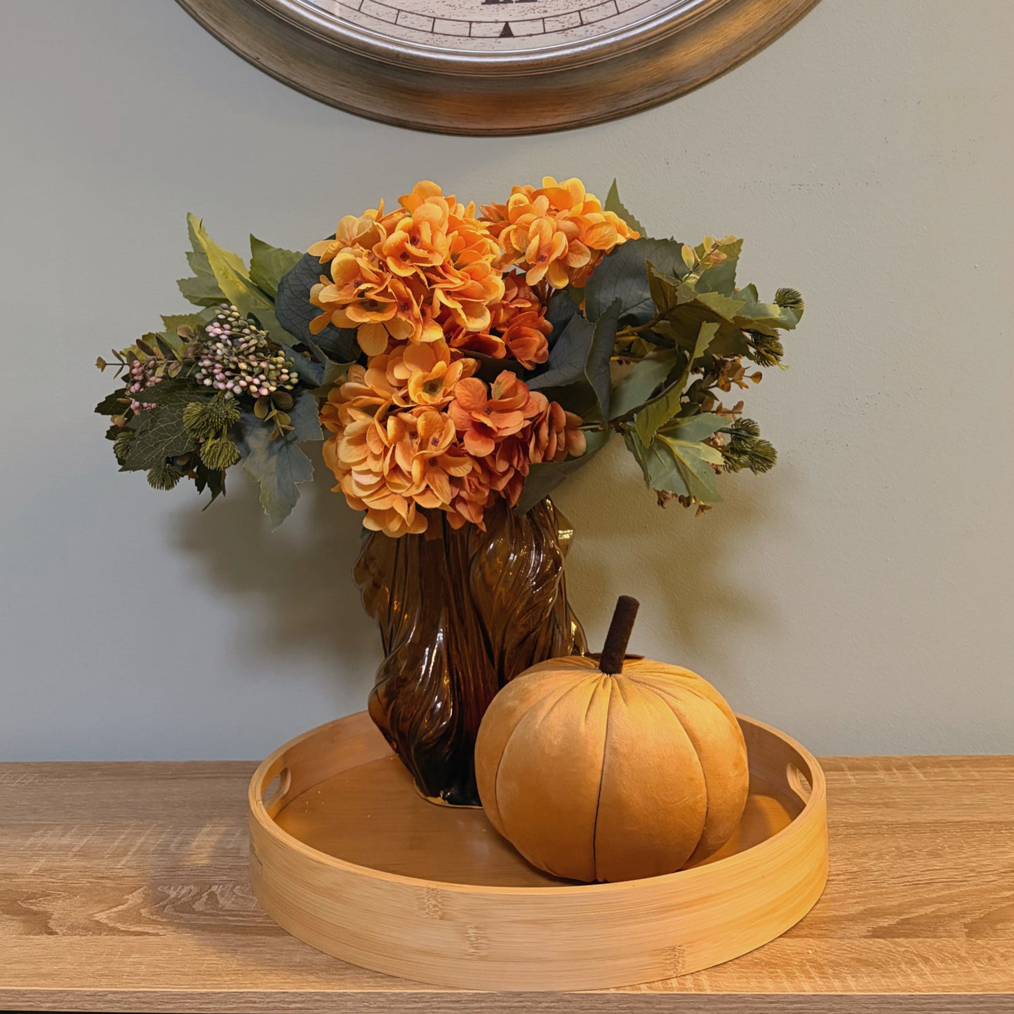 Zucca decorativa in velluto giallo ocra ambientata su vassoio in legno con vaso e fiori autunnali